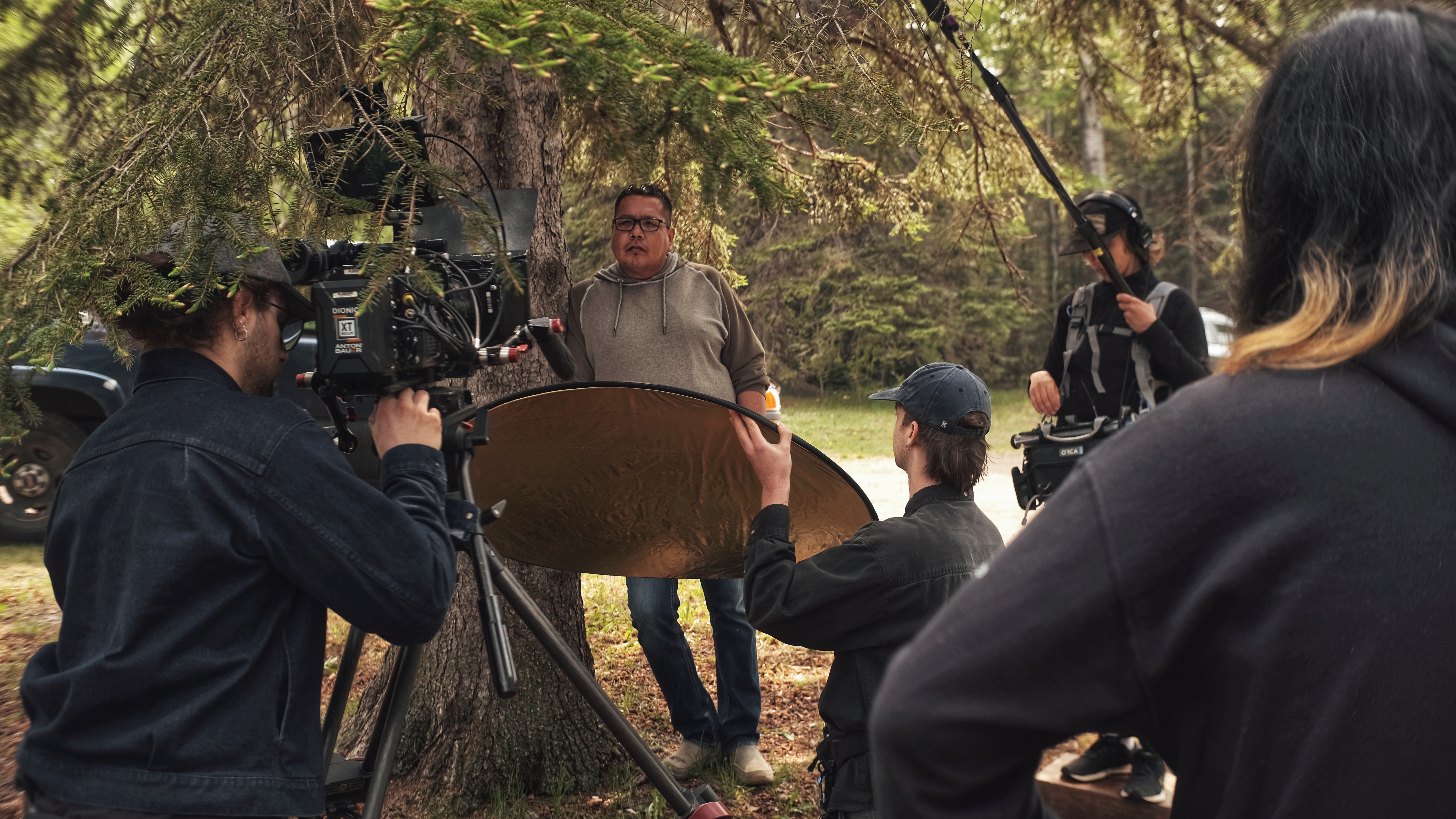 A group of people in the process of creating a film. One person films. Another person holds a light reflector. Another person holds sound recording equipment.